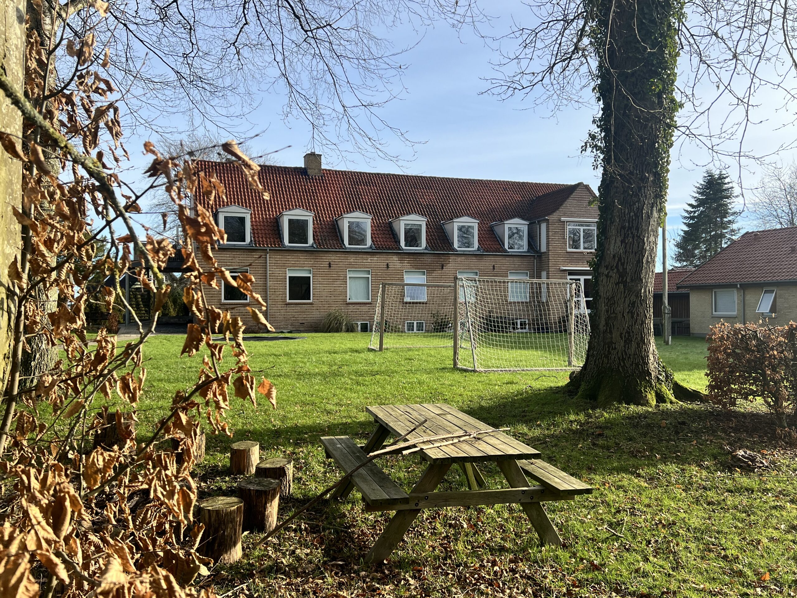 Et slidt træpicnicbord står på en plæne dækket af blade og træstubbe i forgrunden; bagved står et stort træ og to fodboldmål på en solbeskinnet græsplæne foran en lav rødmuret bygning med rødt tegltag og kvistvinduer.