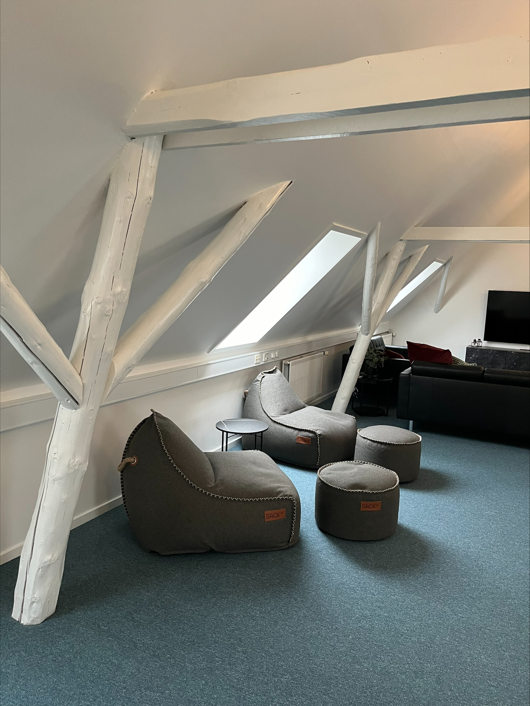 To grå loungestole med lædermærker med teksten u0022SACKu0022 og to runde puffer står på blåt tæppe under et skråt loft med hvide malede bjælker, med et lille rundt sidebord mellem stolene og en sort sofa og tv i baggrunden.