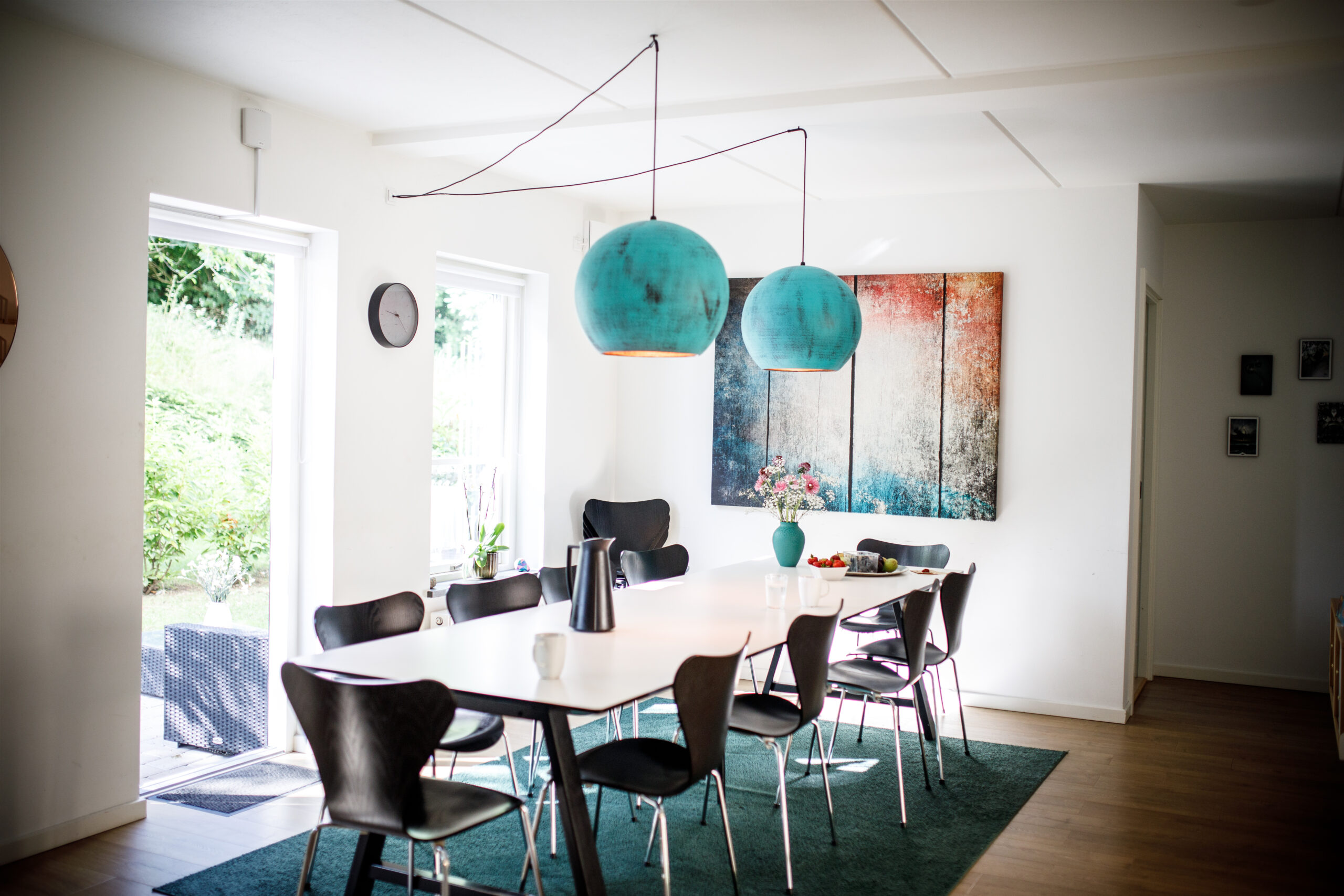 Langt hvidt spisebord omgivet af sorte stole står midt i et lyst spiserum med to store turkise pendellamper hængende over bordet. På bordet står en grøn vase med blomster, en kande og skåle, og et stort abstrakt maleri hænger på væggen bagved.