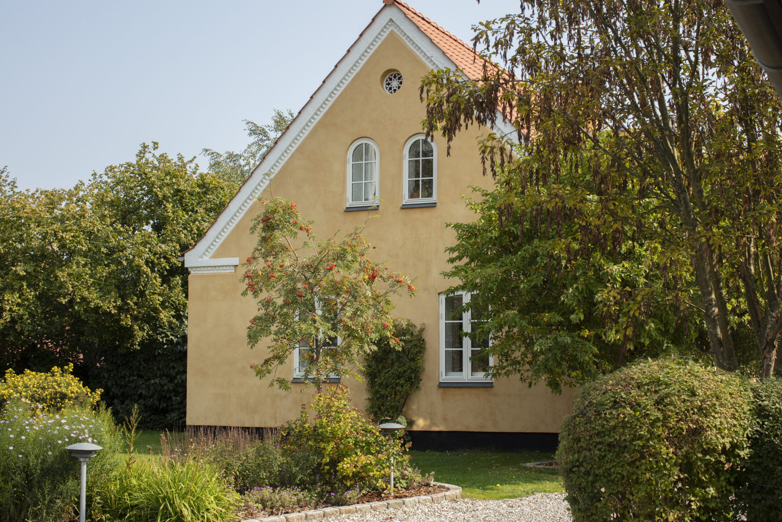Gult pudset hus med rødt tegltag og spids gavl, to buede vinduer under gavlen og en lille rund ventil, set fra en forhave. Forgrunden viser en frodig have med buske, blomsterbede, et grusareal og en lav havelampe.