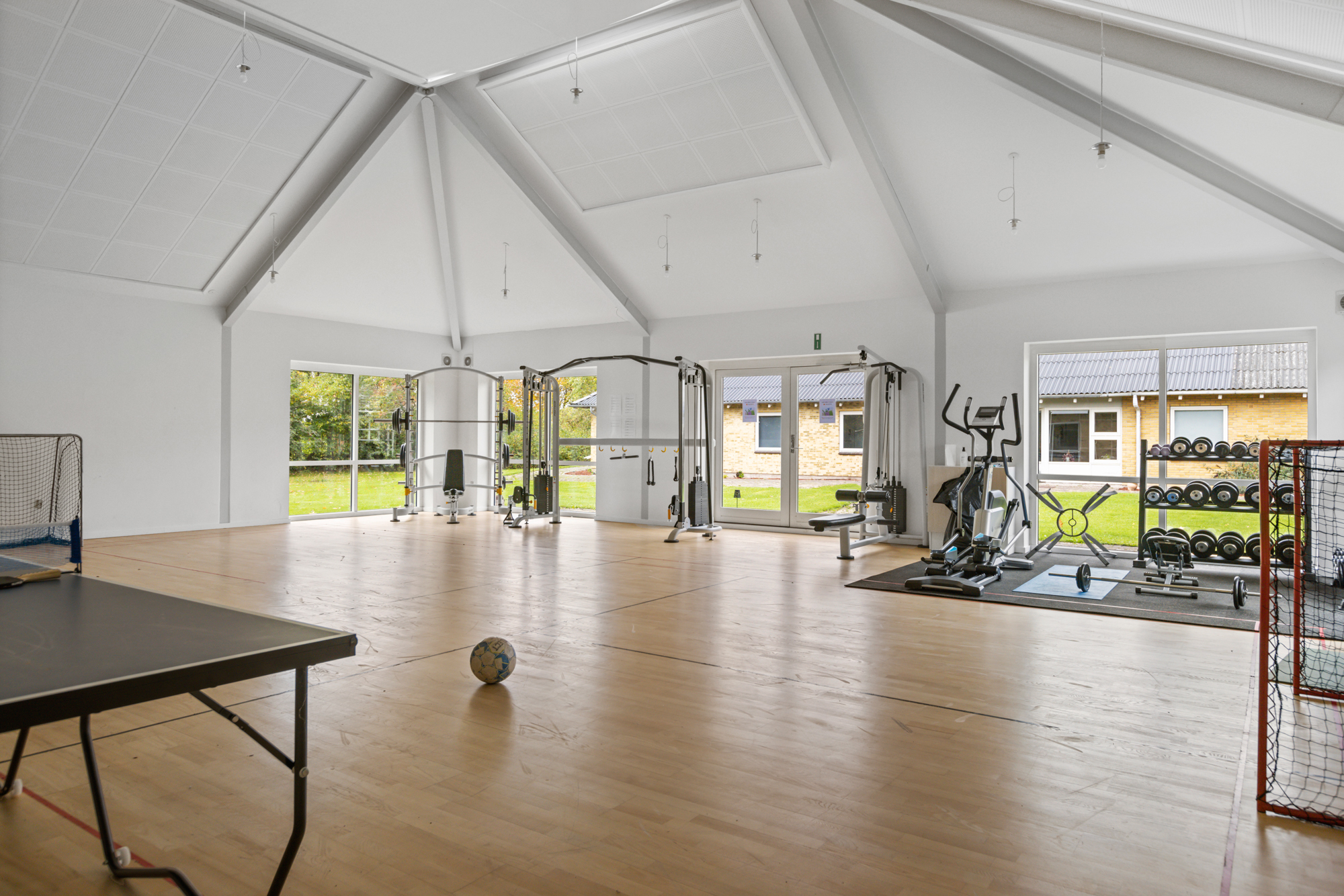 Stor, lyst træningsrum med højt hvidt loft og store vinduer; en bold ligger midt på det polerede trægulv, og rummet rummer et kabeltræningsapparat, konditionsmaskiner, vægtstativ, bordtennisbord og små hockeymål.