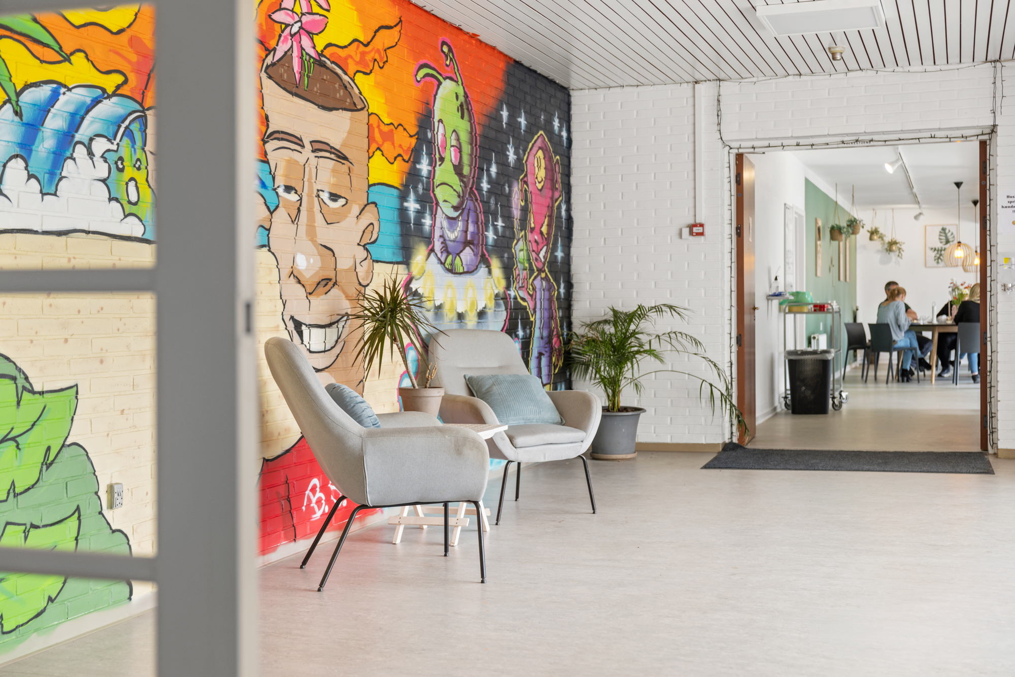 To grå lænestole med puder står foran en farverig graffiti-malet mur i en lys gang; potteplanter står ved stolene, og gennem en åben dør til højre ses et lokale med flere personer siddende ved et bord.