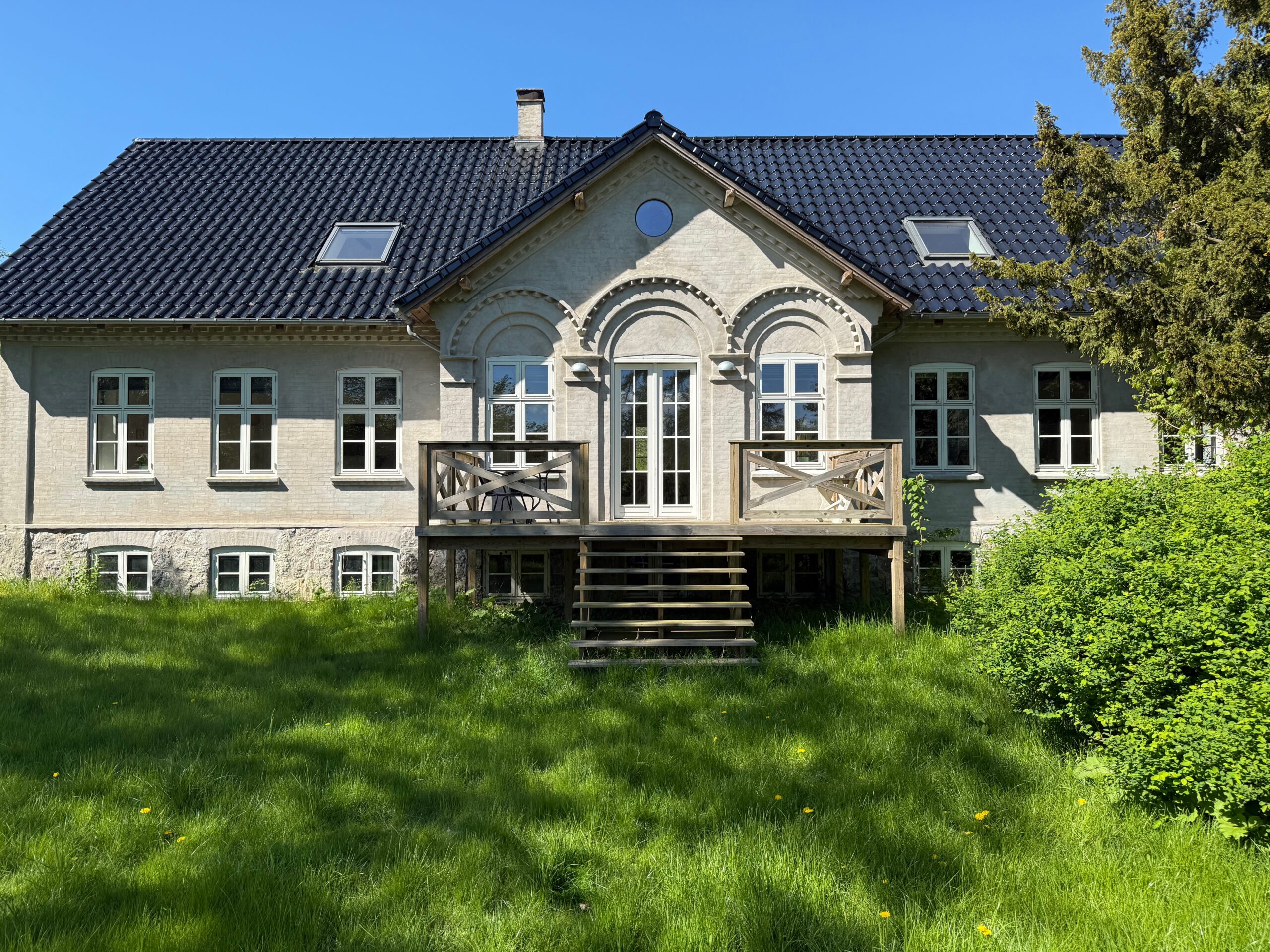 Stort ældre lyst hus med sort tegltag og et centralt gavlparti med dekorative buer; franske døre fører ud til en lille træaltan med trappe ned til en frodig grøn græsplæne, omgivet af buske og en klar blå himmel.