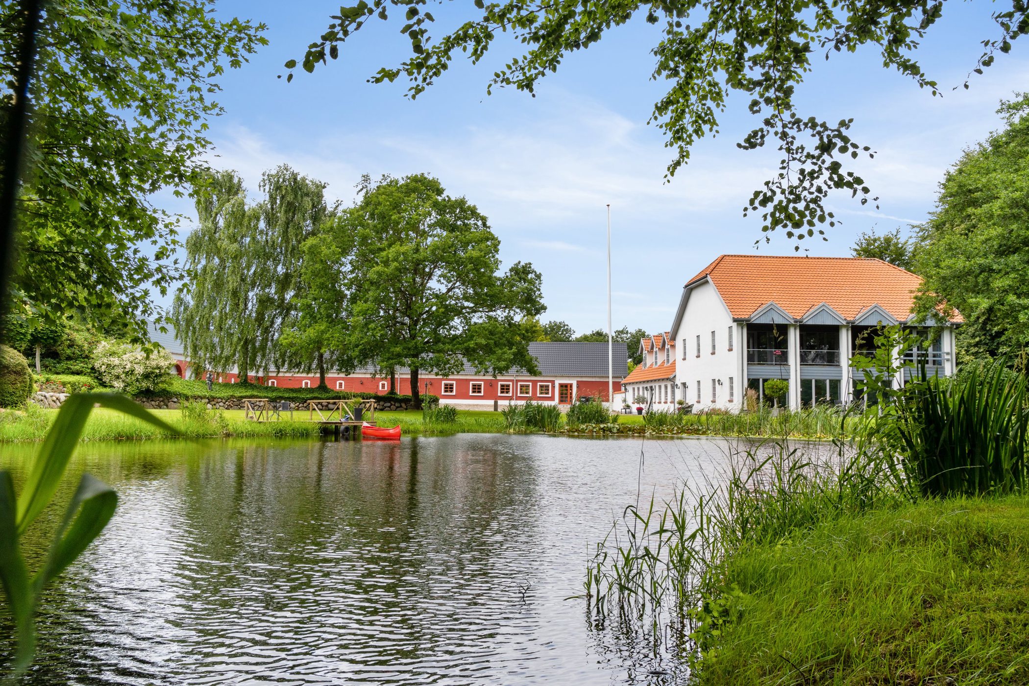 Sø med let bølgende vand i forgrunden omgivet af grønt græs og siv, en rød kano ved en lille træbro; i baggrunden en lang rød bygning og et hvidt hus med orange tegltag og altaner, indrammet af store grønne træer og hængende grene.