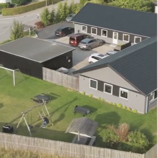 Luftfoto af et lille boligområde med to grå bygninger og en parkeringsplads med flere biler; i forgrunden ses et indhegnet græsareal med et gyngestativ hvor en person sidder, to picnicborde, et overdækket siddeområde og et lille fodboldmål.