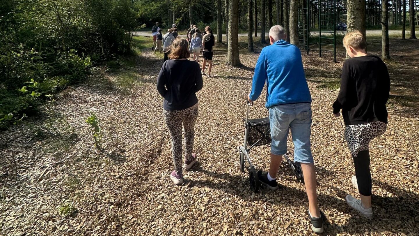 En gruppe mennesker går væk fra kameraet ad en sti dækket af flis i en lys skov; forrest ses tre personer set bagfra, hvoraf den midterste skubber en rollator, og et klatrestativ fra en legeplads er synligt til højre.