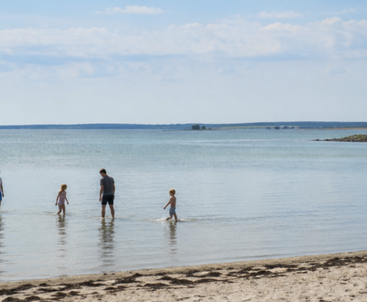 To voksne og to børn vader i lavt kystnært vand tæt på en sandstrand, spredt ud i en linje mod havet. Himmel med lette skyer og roligt vand strækker sig ud mod horisonten.