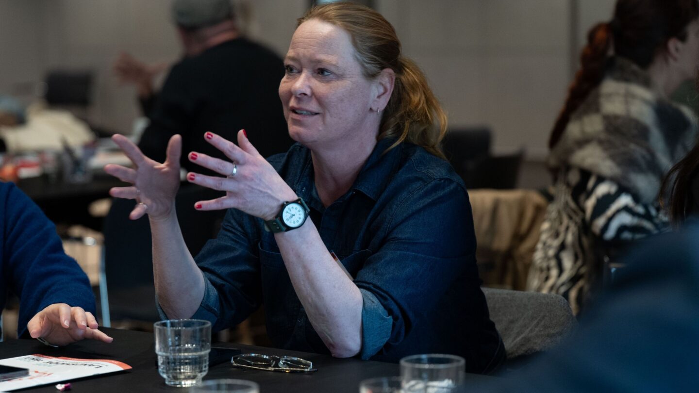 Kvinde med sløret ansigt og opsat hår, iført mørk denimskjorte, gestikulerer med begge hænder over et bord under et møde; på bordet ligger briller, flere vandglas og papirer, mens andre deltagere sidder sløret i baggrunden.